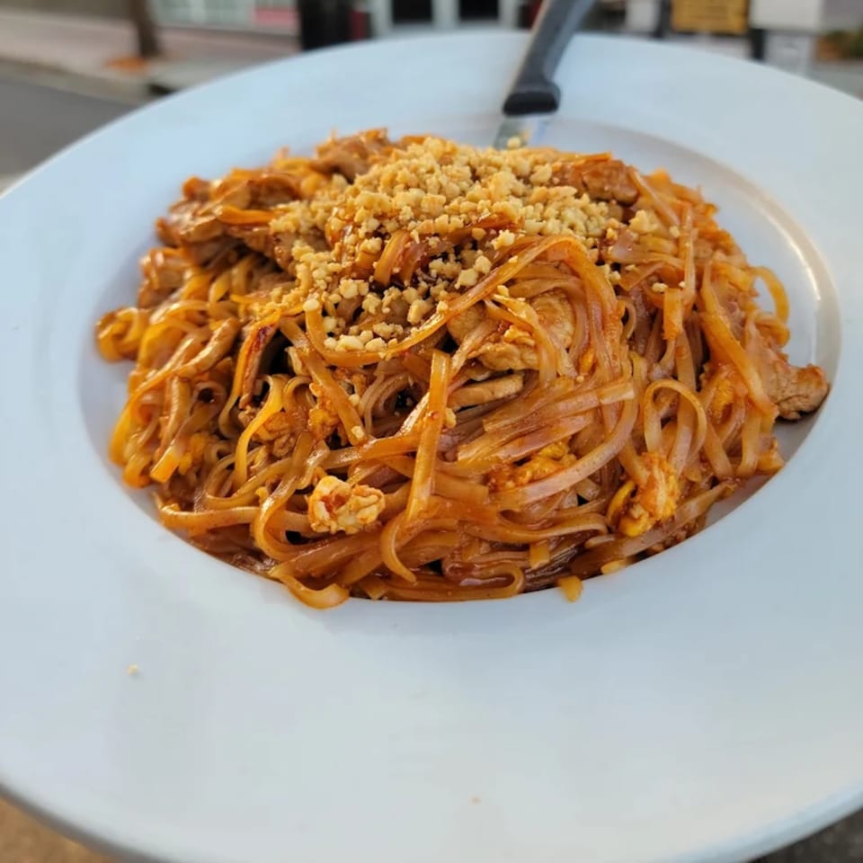 A close-up of a white plate filled with Thai Pad See Ew noodles, topped with crushed peanuts.