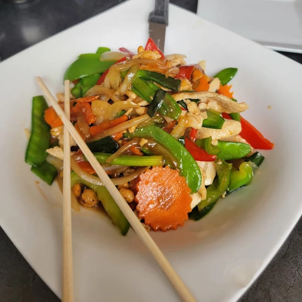 Thai stir-fry with chicken, colorful vegetables, and peanuts, served with chopsticks on a white plate.