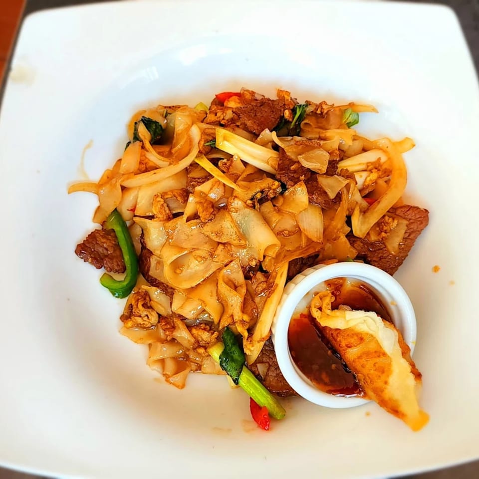 Thai Drunken Noodles with beef, onions, peppers, and a side of dipping sauce with a dumpling.