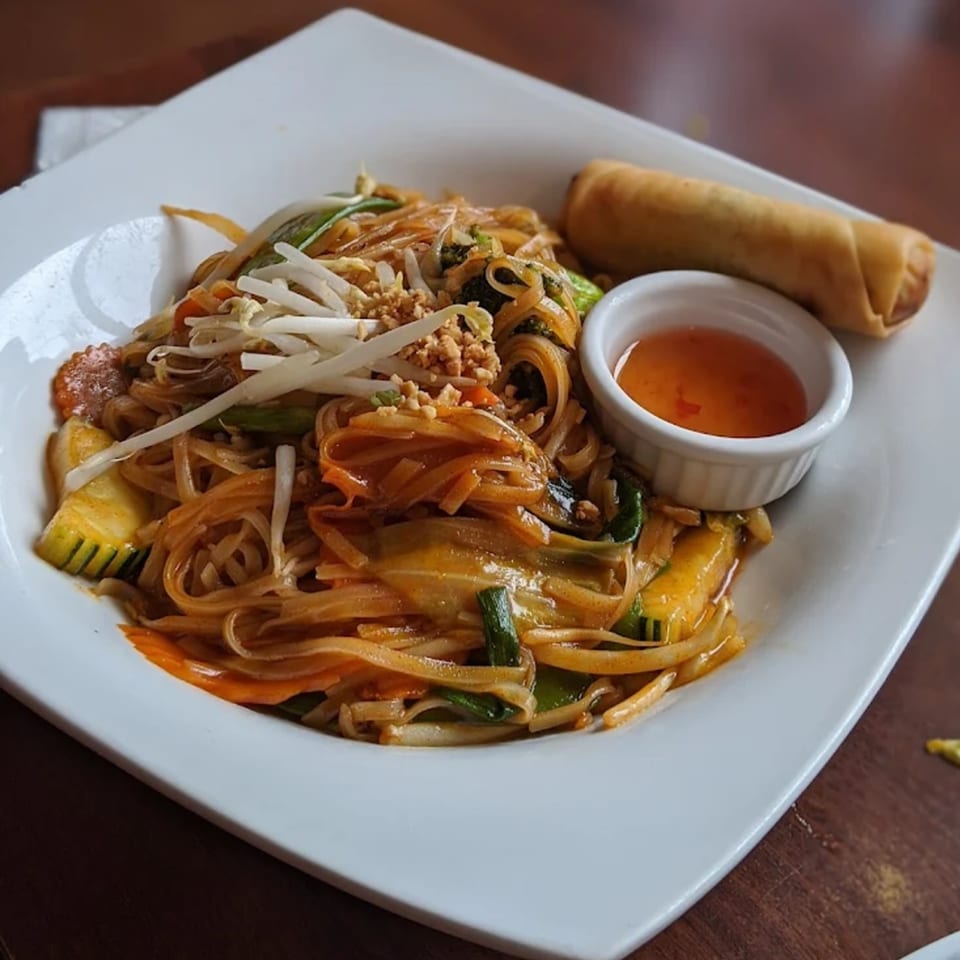 A plate of Thai noodles with vegetables, bean sprouts, crushed peanuts, an egg roll, and dipping sauce.